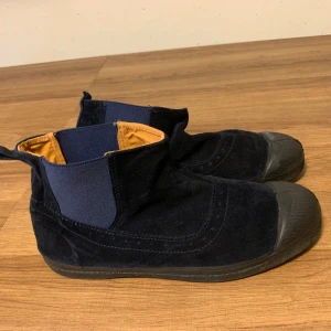 Svarta mocka boots Bensimon tennis - Säljer ett par snygga svarta Bensimon boots i mocka med elastiska paneler på sidorna för enkel på- och avtagning. De har en robust gummisula och tå, vilket gör dem perfekta för höst och vinter. Insidan är fodrad i en kontrasterande orange färg som ger en cool touch. Perfekta för både vardag och lite mer uppklädda tillfällen! Använda 1 gång