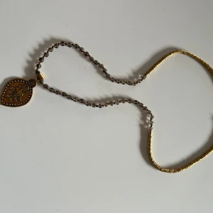 Halsband med guld- och bruna detaljer från Syster P - Unikt halsband med en vacker medaljong i guld och brunt. Kedjan är delvis dekorerad med små pärlor som ger en elegant touch. Perfekt för att lyfta en outfit till fest eller speciella tillfällen.