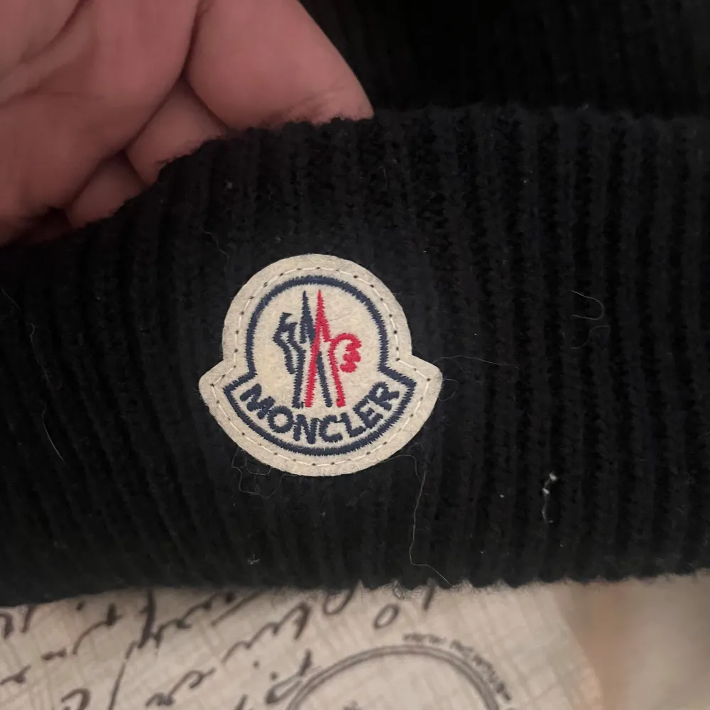 Säljer en snygg svart stickad mössa från Moncler. Den har en klassisk ribbad design och ett broderat Moncler-märke framtill. Perfekt för att hålla sig varm och stilren under kalla dagar. Passar till alla vinteroutfits!. Accessoarer.