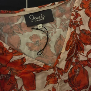 Blommig klänning från Jewels by Indiska - Säljer en superfin blommig klänning från Jewels by Indiska i storlek XS. Den har en v-ringad design och långa ärmar, perfekt för både vår och sommar. Klänningen har ett härligt mönster i rött och vitt som verkligen poppar!