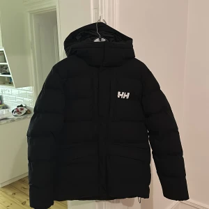 Svart pufferjacka från Helly Hansen - Säljer en svart pufferjacka från Helly Hansen i mycket bra skick. Jackan har en varm och vadderad design med huva och dragkedja framtill. Perfekt för kalla vinterdagar. Helly Hansen-logga syns både fram och bak. Passar perfekt för vinterns alla äventyr! ❄️Inköpt förra året, säljer pga den blivit förstor för mig 