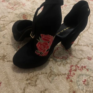 Svarta klackskor med rosbroderi (DinSko) - Snygga svarta klackskor med vackert rött rosbroderi på klacken. De har en elegant rem runt ankeln och en bekväm blockklack. Perfekta för en festlig kväll eller en utekväll med vänner. Skorna är i bra skick och ger en romantisk touch till din outfit.