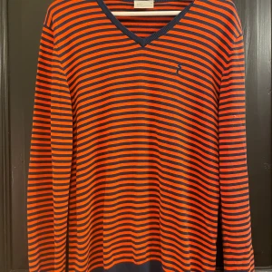 Randig tröja från Peak Performance - Säljer en snygg randig tröja från Peak Performance i storlek M. Den har en cool kombination av orange och mörkblå ränder med en diskret logga på bröstet. Perfekt för både höst och vår. Tröjan är i mycket bra skick och har en bekväm passform med långa ärmar och v-ringning.