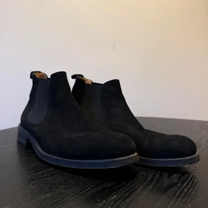 Svarta mocka Chelsea boots - Säljer ett par stilrena svarta Chelsea boots i mocka. De har en klassisk design med elastiska paneler på sidorna för enkel på- och avtagning. Sulan är robust med bra grepp, perfekt för både vardag och fest. Insidan är bekväm och fodrad för extra komfort. Passar perfekt till höst- och vintergarderoben!