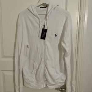 Vit hoodie från Ralph Lauren - Säljer en snygg vit hoodie från Ralph Lauren i storlek M. Den har en dragkedja framtill och en klassisk logga på bröstet. Perfekt för en avslappnad stil och passar till alla tillfällen. Huvan är justerbar med snören och det finns praktiska fickor framtill. Helt ny med tag kvar! Mycke bra skick inga skador