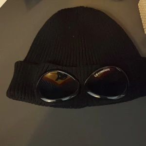 Svart mössa från C.P. Company - Säljer en svart stickad mössa från C.P. Company. Den har en cool design med inbyggda glasögon på framsidan. Perfekt för att hålla sig varm och se stilren ut under kalla dagar. Mössan är i mycket bra skick och passar de flesta tack vare sin one size-storlek.