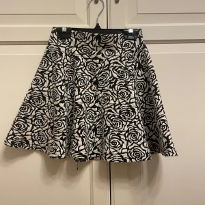 Svartvit kjol med rosor från H&M Divided - Snygg svartvit kjol från H&M Divided med ett iögonfallande ros-mönster. Kjolen har en dragkedja bak och är perfekt för att ge en stilfull touch till din outfit.
