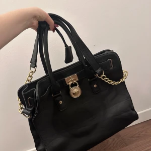 Svart handväska från Michael Kors - Elegant svart handväska i skinn från Michael Kors med guldfärgade detaljer och lås. Väskan har dubbla handtag och en kedjedetalj. Insidan är fodrad med ett ljust tyg med MK-logga och har flera fack för enkel organisering.
