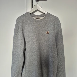 Grå sweatshirt från Moncler - Säljer en stilren grå sweatshirt från Moncler med rund hals och deras ikoniska logga på bröstet. Perfekt för en avslappnad och trendig look.