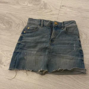 Jeanskjol från Lindex - Säljer en cool jeanskjol från Lindex i storlek 128. Kjolen har en klassisk blå denimfärg med slitna detaljer och rå kant. Perfekt för en avslappnad stil.