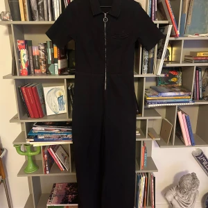 Denim byxdress från Urban Outfitters - Snygg mörkblå denim jumpsuit från Urban Outfitters med korta ärmar och en framträdande dragkedja framtill. Tillverkad i bomull med en liten del elastan för extra komfort. Den är stretchig men med breda ben så den sitter supersnyggt och kan stylas på massa olika sätt, både sexy och edgy på samma gång! 