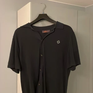 Svart kortärmad skjorta från Morris  - Snygg svart kortärmad skjorta från Morris med knappar framtill och en broderad logga på bröstet. Perfekt för en stilren look.