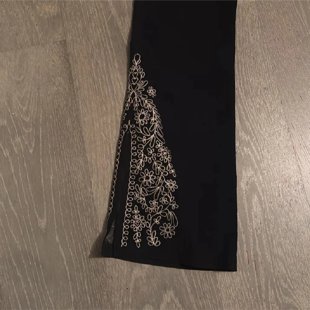 Snygga svarta byxor från Miss Etam med vackra broderade blommor i guld längs benen. Perfekta för en festlig kväll eller när du vill känna dig extra fin. Byxorna har en elegant lågmidjade bootcut-stil mocca tyg som ger en smickrande siluett. De är i mycket bra skick och redo att bäras till nästa event! Midjemått ca 36cm innerbenslängd 83cm. Farkut & Housut.