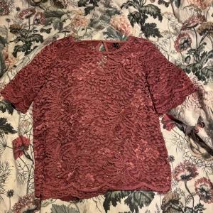 Rostfärgad spetsblus - Säljer en vacker rostfärgad spetsblus med korta ärmar och rund hals. Blusen har ett fint mönster och en knappdetalj i nacken. Perfekt för en elegant look.