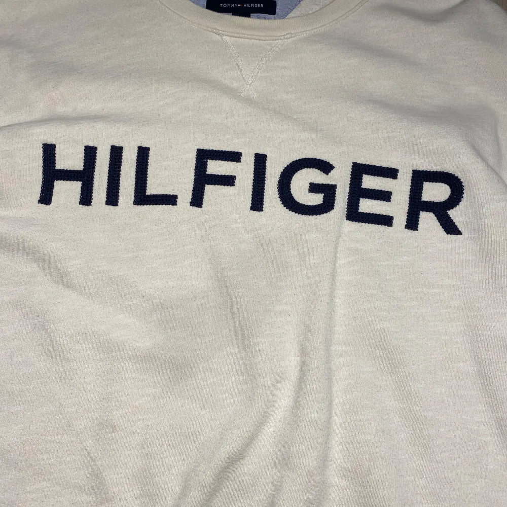 Säljer en stilren vit tröja från Tommy Hilfiger med mörkblå text på framsidan. Tröjan har långa ärmar och en klassisk rund halsringning med ribbade muddar. Perfekt för en avslappnad look.. Neuletakit & Villapaidat.