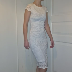 Vit spetsklänning - Elegant vit klänning i spets med korta ärmar och figurnära passform. Klänningen har en vacker spetsdetalj vid fållen och en rund halsringning. Perfekt för sommarens festligheter. Köpt second hand, aldrig använd av mig. Nyskick förutom lite gula under armarna men det är inget som syns när klänningen är på. Passar xs-m