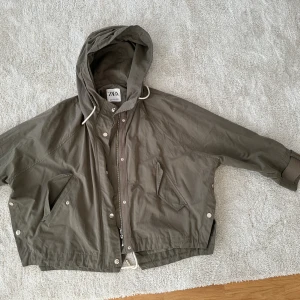 Olivgrön vindjacka från ZARA - Snygg olivgrön vindjacka från ZARA med huva och dragkedja. Jackan har en avslappnad passform och praktiska fickor framtill. Perfekt för blåsiga dagar.
