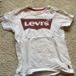 Vit t-shirt från Levi's - Säljer en klassisk vit t-shirt från Levi's med det ikoniska röda logotyptrycket på bröstet. T-shirten är kortärmad och har en rund halsringning innanför med röd detalj. Perfekt för en avslappnad stil💕