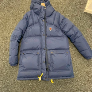 Blå dunjacka från Fjällräven - Säljer en blå dunjacka från Fjällräven med huva och gula detaljer. Jackan har långa ärmar och praktiska fickor med knappar. Perfekt för kalla dagar. Inte använd mer än 2 gånger säljer nu på grund av flytt 