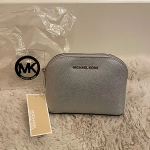 Michael Kors Cindy necessär i silver  - Superfin necessär från Michael Kors. Modellen heter ”Cindy travel pouch”. Silverfärgat läder samt silver detaljer. Har gott om utrymme inuti och en liten ficka. Är helt ny och oanvänd, kommer med prislappen kvar, skydd om dragkedjan, MK care card och plastpåse. Har dock en liten fläck nära dragkedjan, men inget som syns om man inte kollar nogranna (se bild 4). Mått ca 17x13x6,5 cm. 💕
