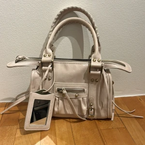Beige handväska med spegel - Snygg beige handväska med nitar. Väskan har två handtag och en praktisk spegel som hänger på framsidan. Perfekt för att hålla dina saker organiserade och stiliga.   Bredd:  21cm   Höjd:  18   💋💋💋