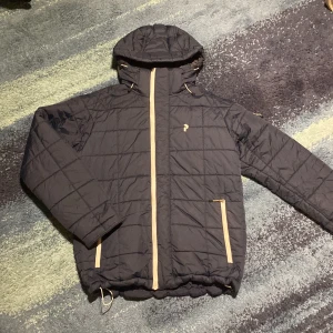 Blå peak performance jacka - Säljer en blå  quiltad jacka från Peak Performance med huva och dragkedja. Perfekt för kyligare dagar med sin stilrena design och praktiska fickor. Jackan har en diskret logga på bröstet och är tillverkad i slitstarkt material. Nästan helt oanvänd skick 9/10