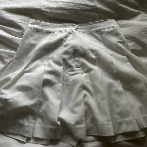 Vit kjol från Divided - Snygg vit kjol från Divided med veckad design. Perfekt för en stilren look. Kjolen har en knapp framtill och är i en kort modell.Jag skulle gissa att jag köpt kjolen på H&M. Använd max 1 gång. 🫶🏻