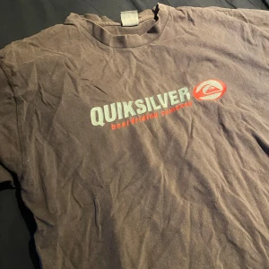 Brun t-shirt från Quiksilver - Säljer en brun t-shirt från Quiksilver med tryck i silver och rött på framsidan. T-shirten är kortärmad och har en klassisk rund halsringning. Perfekt för en avslappnad stil.