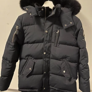 Svart pufferjacka från Moose Knucklers - Säljer en mörkblå pufferjacka från Moose Knucklers med avtagbar huva och pälsdetaljer. Jackan har flera fickor med knappar samt ett broderat motiv på insidan av jackan. Perfekt för väldigt kalla dagar!
