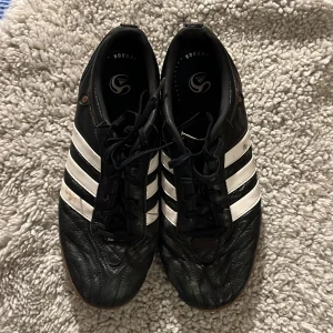 Svarta Adidas fotbollsskor - Säljer ett par svarta Adidas fotbollsskor med klassiska vita ränder. Skorna har snörning och en slitstark sula, perfekt för inomhusspel. De är i syntetmaterial och har en sportig look. Obs: hål i sidan! 