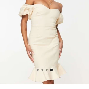 Beige offshoulder klänning - Elegant beige offshoulder klänning med volangdetaljer vid fållen. Klänningen har korta puffärmar och en figurnära passform. Perfekt för en stilfull kväll. Aldrig använd. Nypris 829:-