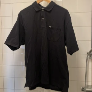 Svart pikétröja från Yoke - Snygg svart pikétröja från Yoke med krage och knappar. Tröjan har en bröstficka med en liten Yoke-logga. Perfekt för en stilren look.