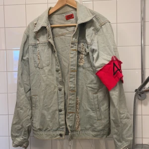 Grå jeansjacka från 424 - Säljer en grå jeansjacka från 424 med slitna detaljer och en röd armbindel. Jackan har knappar framtill och två bröstfickor. Perfekt för en edgy stil!