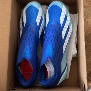 Adidas X Crazyfast.1 FG Fotbollsskor - Säljer ett par Adidas X Crazyfast.1 FG fotbollsskor i en snygg blå färg med vita detaljer. Skorna har en modern design med en lätt och smidig konstruktion, perfekt för snabba rörelser på planen. Sulan är utrustad med dobbar för optimalt grepp.