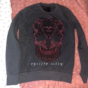 Grå tröja med dödskalle från Philipp Plein - Snygg grå tröja från Philipp Plein med ett coolt dödskallemotiv i svart och rött på framsidan. Tröjan har långa ärmar och rund halsringning. Perfekt för en edgy look!