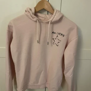 Rosa hoodie från Gina Tricot - Säljer en söt rosa hoodie från Gina Tricot med texten 'Ma Chérie' och en stjärna på bröstet. Tröjan har en mysig huva med dragsko och långa ärmar. Perfekt för en avslappnad stil.