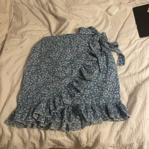 Blå blommig kjol med volang - Charmig blå kjol med blommigt mönster och volangdetaljer. Kjolen har en omlott-design med knytband i midjan, vilket ger en feminin touch. Perfekt för sommarens varma dagar.