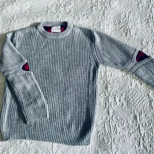 Grå stickad tröja från Zara Kids - Säljer en grå stickad tröja från Zara Kids med coola röda lappar på armbågarna. Tröjan har en klassisk rund hals och långa ärmar. Perfekt för kyliga dagar!