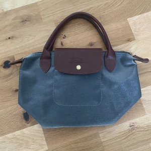 Blå och brun handväska från Longchamp - Snygg handväska från Longchamp i en stilren blå färg med bruna skinnhandtag och detaljer. Väskan har en praktisk dragkedja och en knappstängning framtill. Perfekt för att bära dina dagliga nödvändigheter med stil.