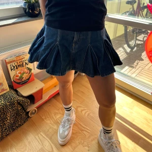 Jeanskjol med volang - Trendig jeanskjol med volangdetaljer. Kjolen har en kort längd och en mörkblå färg som passar perfekt för en avslappnad stil. Perfekt för att ge din outfit en cool touch.