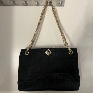 Svart quiltad axelväska med guldkedja - Elegant svart axelväska med quiltat mönster och guldfärgade kedjehandtag. Väskan har en dekorativ guldfärgad detalj på framsidan och är perfekt för att ge en lyxig touch till din outfit. Perfekt som en lätt medelstor vardags väska. Enkel att stänga med en guldfärgad knapp som man trycker in i hålet och vrider. 