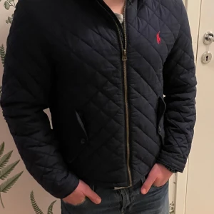 Mörkblå quiltad jacka från Ralph Lauren - Snygg mörkblå quiltad jacka från Ralph Lauren med röd broderad logotyp på bröstet. Jackan har dragkedja och långa ärmar. Klassisk och stilren design som passar till många tillfällen. Storlek xs i storlekn men L i barn storlek