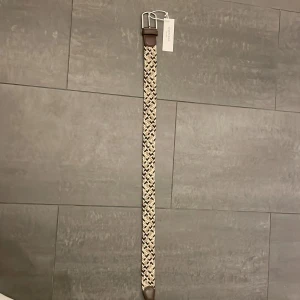 Oanvänt Flätat bälte från Onewear - Snyggt flätat bälte från Onewear i beige och brunt med silverfärgat spänne. Perfekt för att ge en stilren touch till din outfit. Bältet har detaljer i brunt skinn vid spännet och ändarna. Aldrig använd. Med tagg.