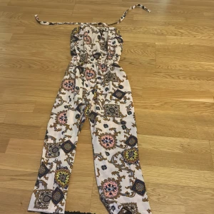 Mönstrad byxdress från Stockholmsklädmärke - Trendig byxdress med ett färgglatt och detaljerat mönster i olika nyanser. Den har en halterneck-design med knytband och en elastisk midja för en bekväm passform. Perfekt för en somrig look!