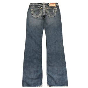 Blå bootcut jeans från True Religion - Snygga blå bootcut jeans från True Religion med ikoniska bakfickor och kontrastsömmar. Perfekta för en avslappnad stil med en touch av retro. Märkeslappen på baksidan ger en extra cool detalj. Kom privat för egna bilder men kan tyvärr inte visa bild på. Midjemåttet rakt över är 40 cm och innerbenslängden är 83 cm🥰