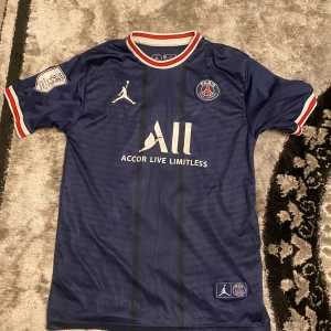 Blå PSG fotbollströja med Mbappé 7 - Snygg blå PSG fotbollströja med korta ärmar och Mbappé 7 tryckt på ryggen. Tröjan har vita och röda detaljer vid krage och ärmslut samt Jordan-logga på framsidan. Perfekt för fotbollsfans! Storlek 176/s