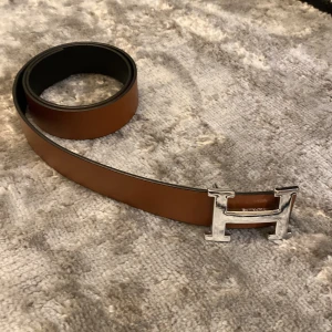 Brunt skärp med silverfärgat H-spänne - Snyggt brunt skärp med ett iögonfallande silverfärgat H-spänne. Perfekt för att ge en stilren touch till din outfit. Skärpet är tillverkat i skinn och har en klassisk design.