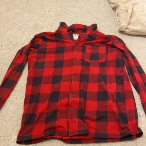 Röd och svart rutig pyjamas skjorta från Kappahl - Snygg röd och svart rutig pyjamas skjorta från Kappahl med långa ärmar och knappar framtill. Skjortan har en bröstficka och är perfekt för en avslappnad stil. 