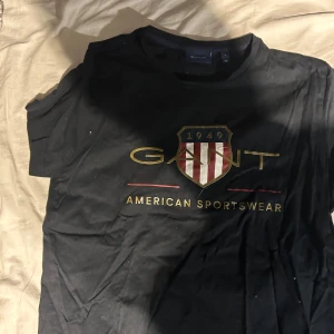 Svart t-shirt från GANT - Snygg svart t-shirt från GANT med tryck av deras logga och texten 'American Sportswear'. Perfekt för en casual look med en touch av stil.Skick 10/10 säljer den på grund av att den aldrig använda pris kan diskuteras vid en snabb affär