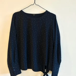 Mörkblå prickig tröja från Opus - Säljer en stilren mörkblå tröja från Opus med små vita prickar. Tröjan har långa ärmar med skortknäopning och motveck. En knytning framtill på sidan ger tröjan en snygg detalj. Perfekt för en chic, lite mer dressad  vardagslook!